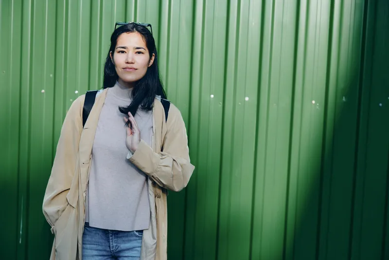 Woman in a light beige coat and grey sweater stands against a vibrant green metal wall, posing with a relaxed hand gesture. Casual street style, layered outfit, urban vibe for lifestyle editorial use