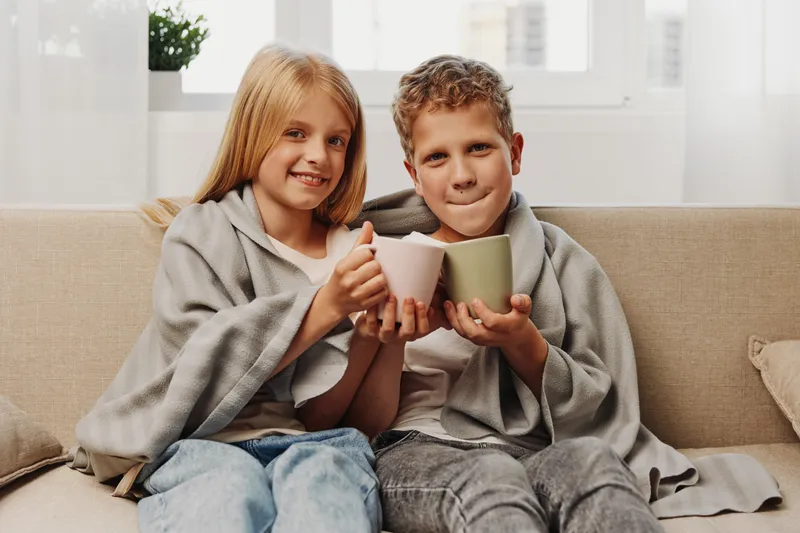 Happy children wrapped in blankets sit on a sofa holding ceramic mugs with warm drinks. Cozy home atmosphere with natural light in the background. Concept of comfort and family time.