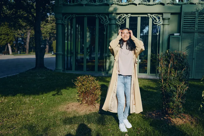 Woman coat outdoors garden casual fashion jeans sneakers smiling sunlight park setting, relaxed pose highlights comfortable style in a natural green landscape with trees and lawn, candid street