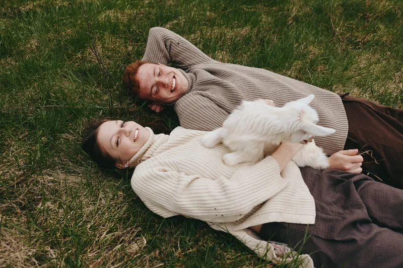 Relaxed outdoor scene of a couple lying on grass with a goat kid, showcasing lifestyle, companionship, and warmth. Soft sweaters, natural light, and nature setting convey comfort and happiness.