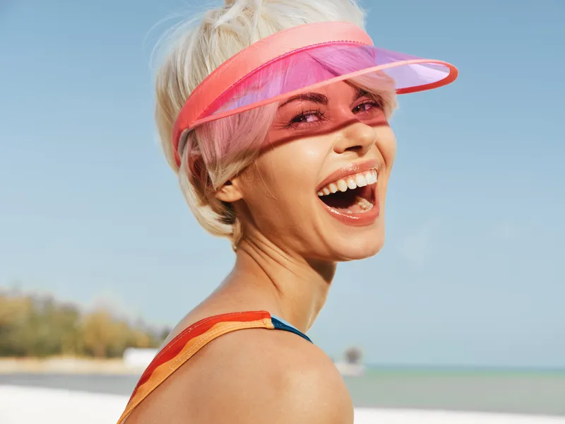 Bright, carefree portrait of a smiling woman at the beach wearing a translucent pink visor, sunlit hair, and a colorful strap, expressing summer happiness and energy.