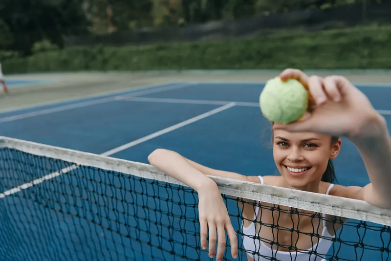 Young female tennis player smiling and holding a tennis ball at the net, showcasing joy and energy, ideal for sports and fitness themes
