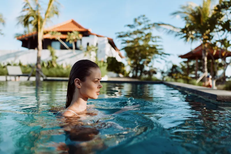 Relaxed woman stands in a sunlit pool with clear water and tropical backdrop, vacation mood, casual swimwear, gentle breeze, peaceful reflection, vibrant colors, resort atmosphere, outdoor leisure