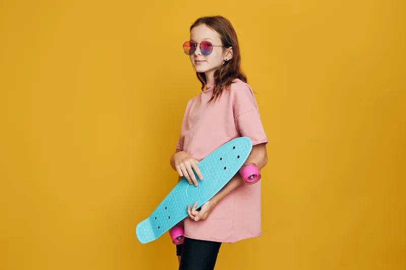Young girl in pink shirt holds blue skateboard with pink wheels posing on yellow background. She wears round sunglasses and looks confident. Casual urban style concept.