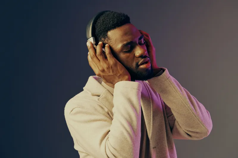A young Black man enjoying music with headphones, dressed in a stylish beige coat, showcasing a relaxed expression against a gradient purple background