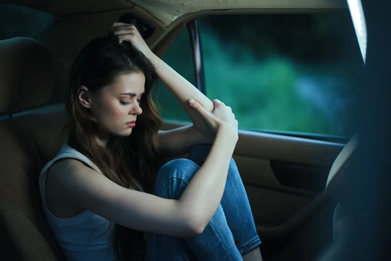 Sad young woman sitting in a car, reflecting on her emotions, wearing casual jeans and a tank top, in a moody, greenish background at dusk.