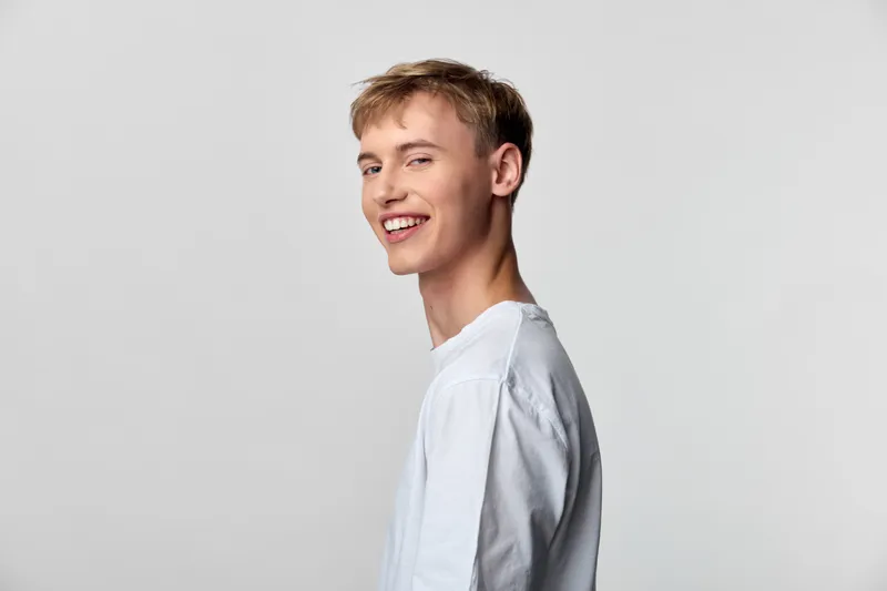 Smile, young man in casual shirt, side view against light backdrop, exuding friendly energy, confident vibe and approachable mood. Clean studio setting emphasizes natural charm and relaxed style.