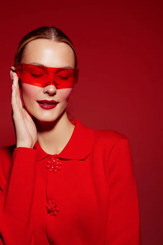 Young woman in red sweater and red translucent glasses poses with eyes closed on red background. Concept of fashion, style, and monochrome portrait with vibrant color.