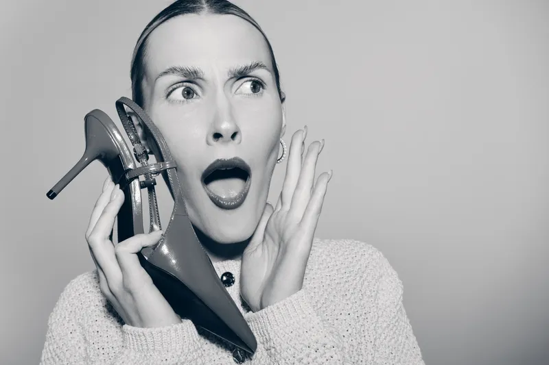 Surprised white woman with light skin holds high heel shoe near ear like a phone, wearing knitted sweater. Concept of shock, fashion, and communication.