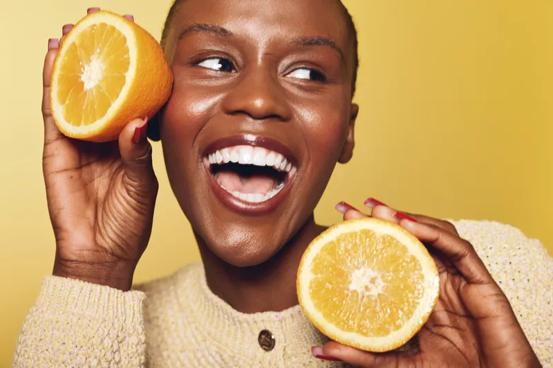 Happy African American woman with dark skin holds two halves of an orange fruit near her face smiling. Bright yellow background and cozy beige sweater create fresh, joyful mood.
