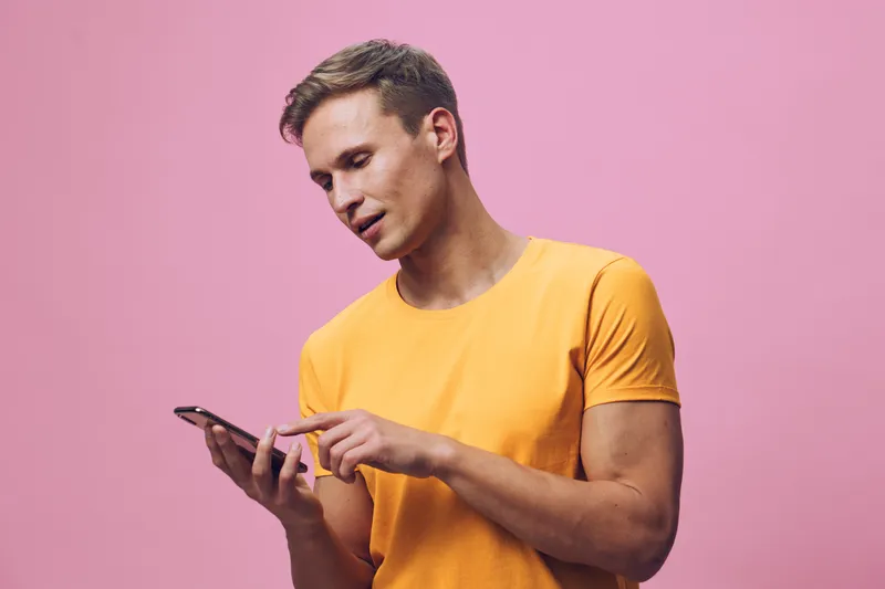 Smiling man wearing yellow t-shirt using mobile phone, looking at screen, isolated on pink background, casual lifestyle shot, popular communication, technology concept, young adult male.