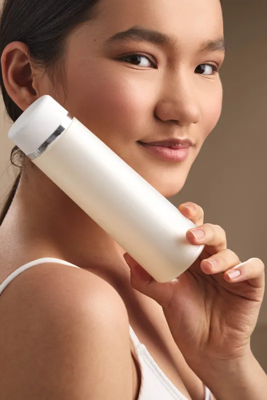 asian woman holds a white cosmetic bottle near her face in a soft studio setting, presenting a skincare moment with calm confidence and a gentle smile that suggests focus and care.