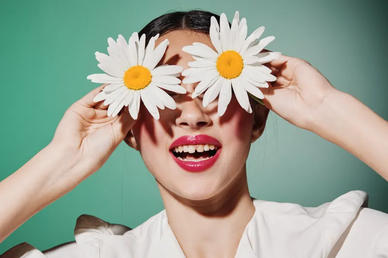 Smiling young woman holding two large daisies over her eyes. The vibrant flower petals contrast with her bright lipstick. Concept of beauty creativity and fashion playfulness.
