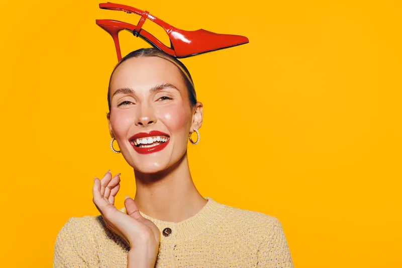 Smiling woman with red lipstick and hoop earrings poses with a red high heel shoe on her head against a bright yellow background. Concept of fashion, fun, and creativity.