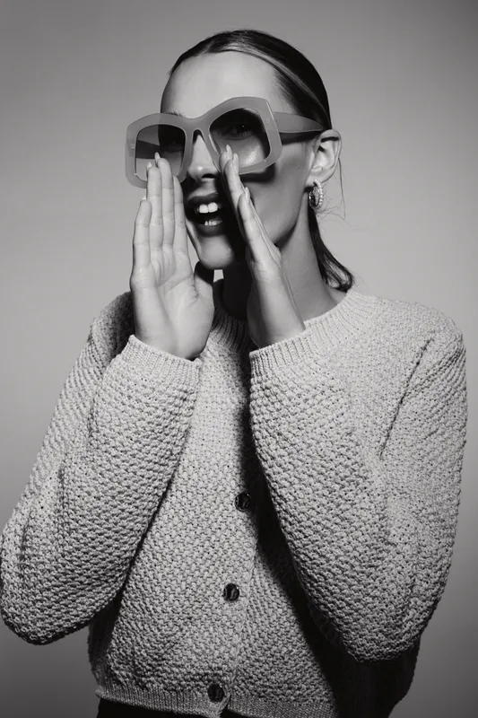 Young white woman with light skin wears knitted sweater and big sunglasses, hands near mouth. Modern fashion and expression concept.