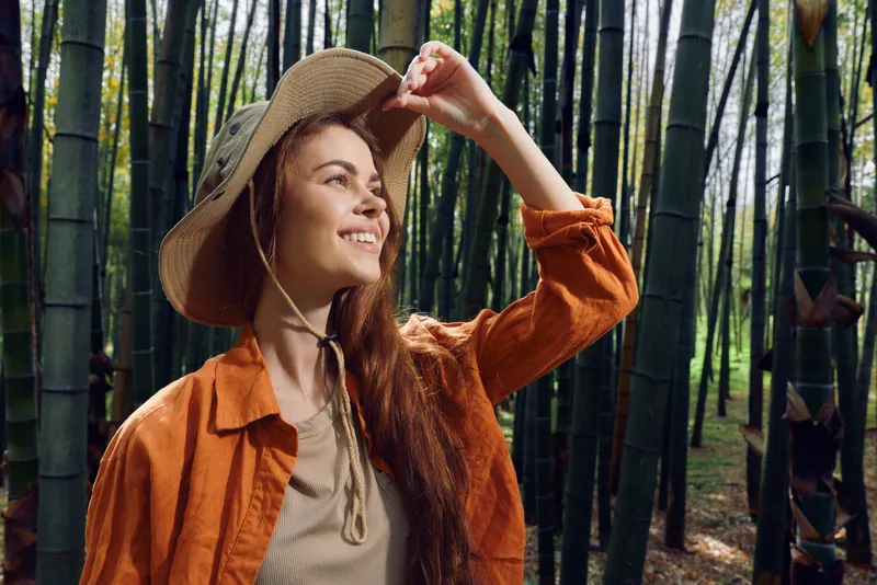 Woman smiling in straw hat standing in dense bamboo forest, nature portrait with warm sunlight on face, travel and eco lifestyle, casual orange jacket and relaxed outdoor mood.