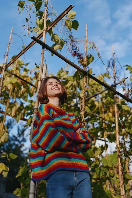Woman in a colorful striped sweater smiling with arms crossed under a garden trellis, short-haired young adult looking up at blue sky, casual outfit and relaxed outdoor lifestyle.