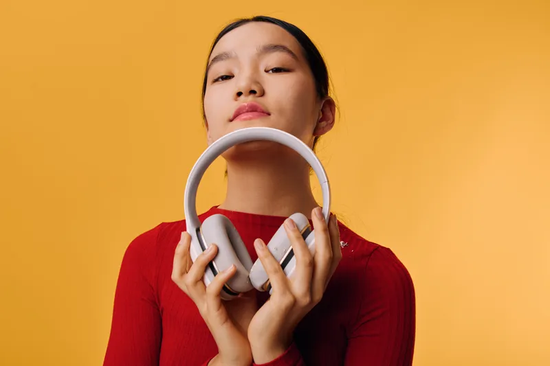 Young woman holds gray headphones close to face with closed eyes against orange background. Relaxed female with earphones in casual outfit for music listening or audio entertainment indoors.