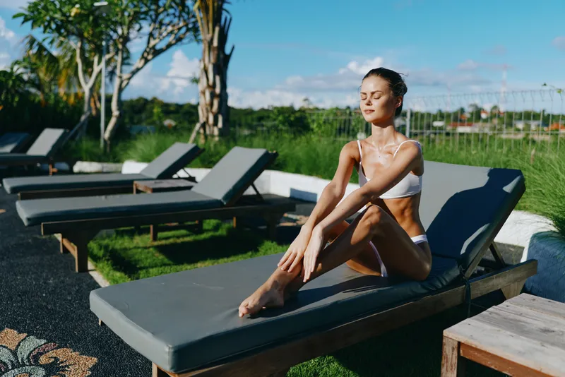 Woman in a white bikini sits on a lounge by a sunny pool, enjoying a relaxed lifestyle outdoors and warm weather, with a tranquil vibe and modern resort atmosphere during daytime.