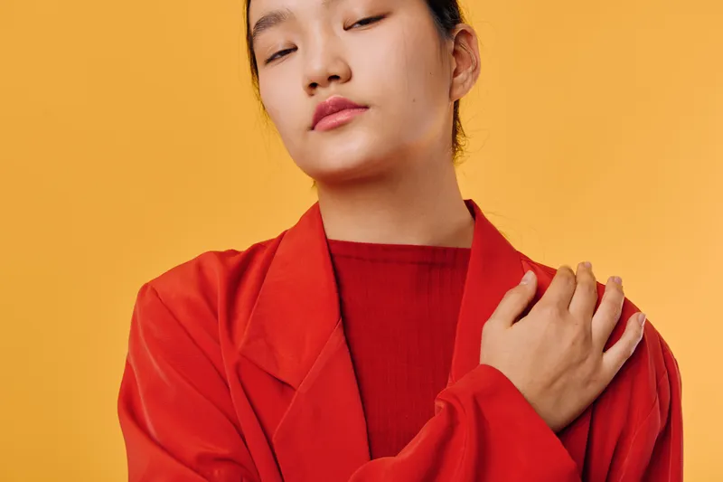 Young woman with closed eyes wearing red jacket and sweater posing with hand on shoulder against yellow background in studio portrait for fashion and emotion.
