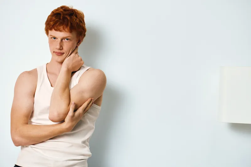 Young man with red hair in white tank top standing near white wall looking at camera with thoughtful expression in casual indoor setting with soft lighting and minimal background.