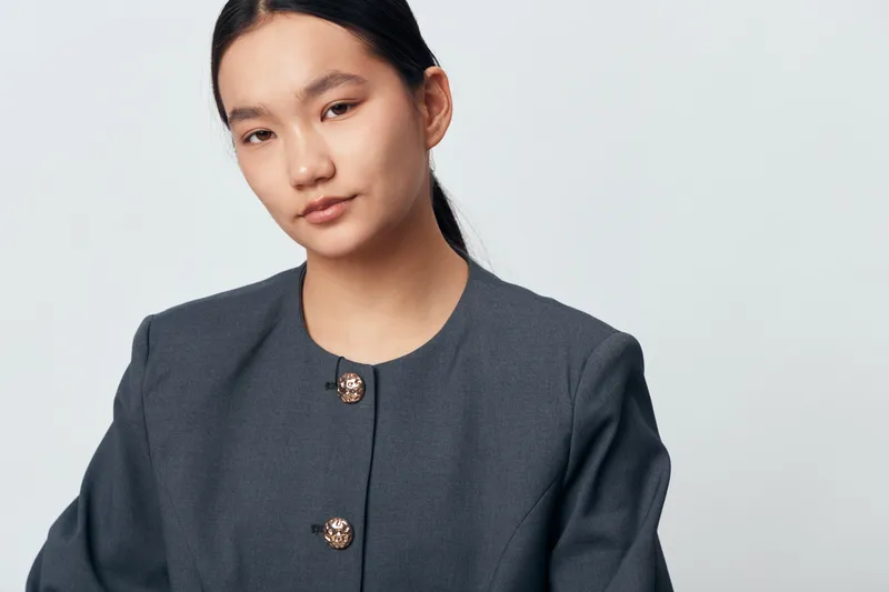 Portrait of young woman with long dark hair wearing gray blazer with decorative buttons standing against light background in studio. Female model with confident and calm expression.