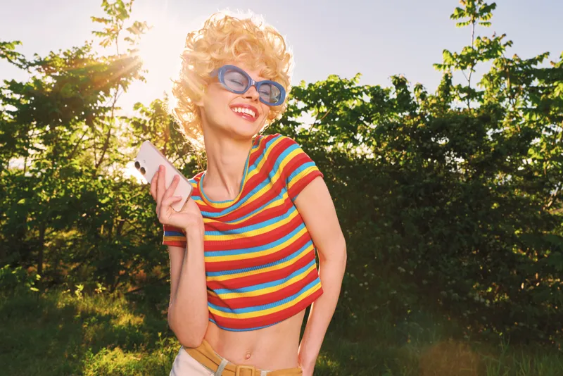 Young woman wearing a colorful striped shirt smiles while checking her phone in bright outdoor light, conveying a playful, carefree mood and a casual lifestyle.