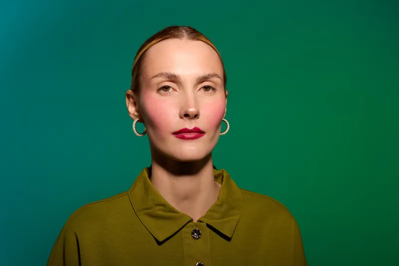 Portrait of a serious woman with red lipstick and hoop earrings wearing a green shirt against a green background. Studio lighting highlights her facial features and expression.