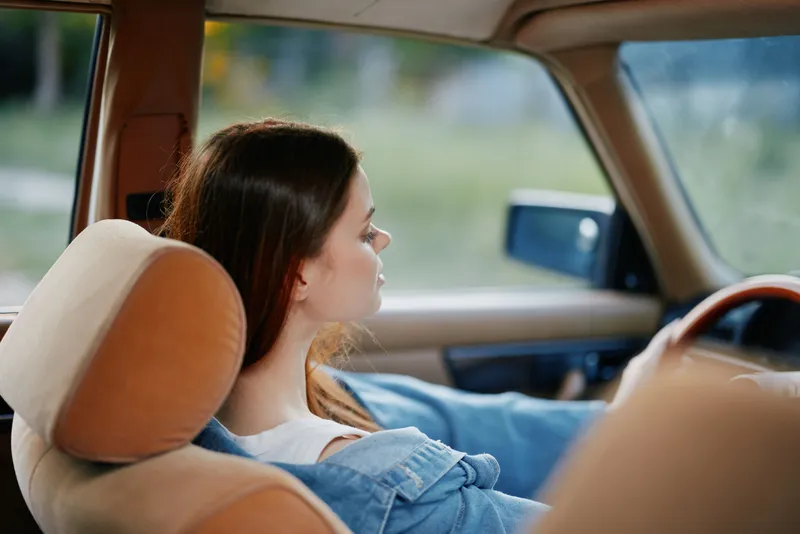 Young woman driving in a vintage car, showcasing serene expression and casual attire in a soft-focus natural background.