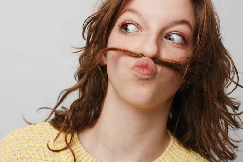 Playful young woman with curly brown hair makes a mustache with her hair, looking sideways with a funny expression. Casual yellow sweater and neutral background create a light mood.