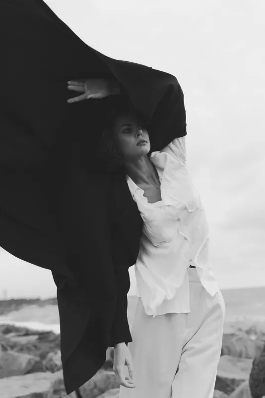 Woman in flowing dress beneath a dark cape on a windy beach, creating a dramatic monochrome silhouette and dynamic movement for editorial stock purposes