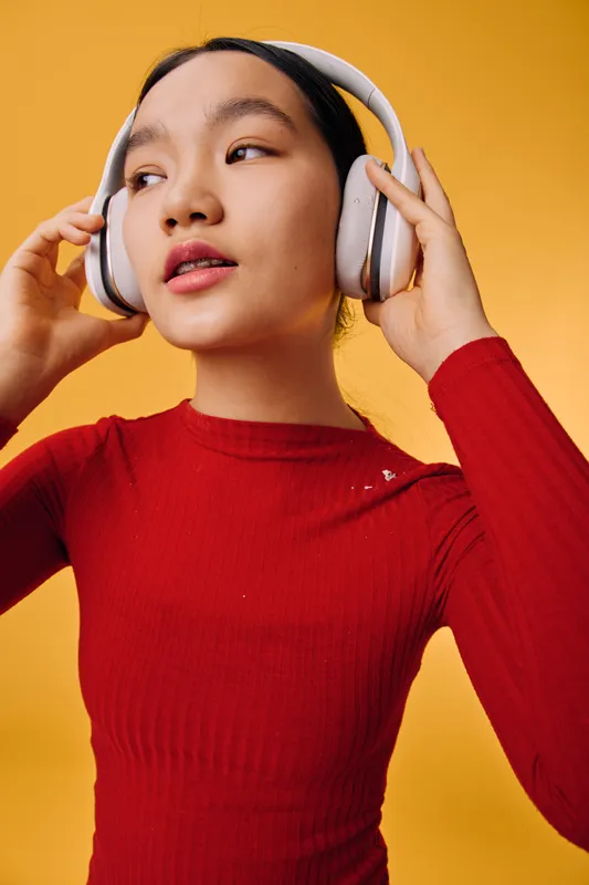 Young woman wearing white headphones listening to music with eyes looking aside. Female in red long sleeve top standing against orange background indoors.