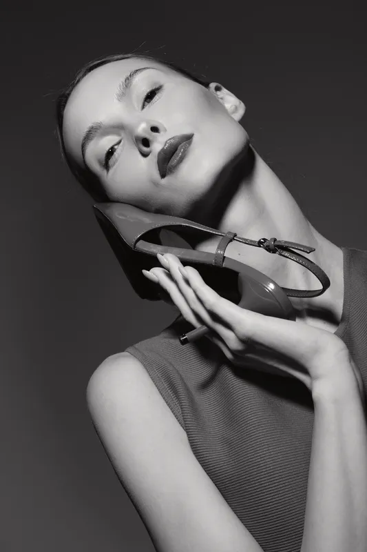 Elegant white woman with pale skin holds high heel shoe near face in black and white studio portrait. Concept of fashion, style, beauty, and glamour with minimalistic lighting.