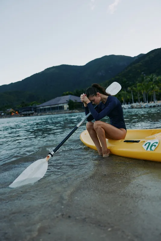 Young woman resting in a kayak on the shore, wearing a navy blue outfit with mountains and water in the background during calm weather.