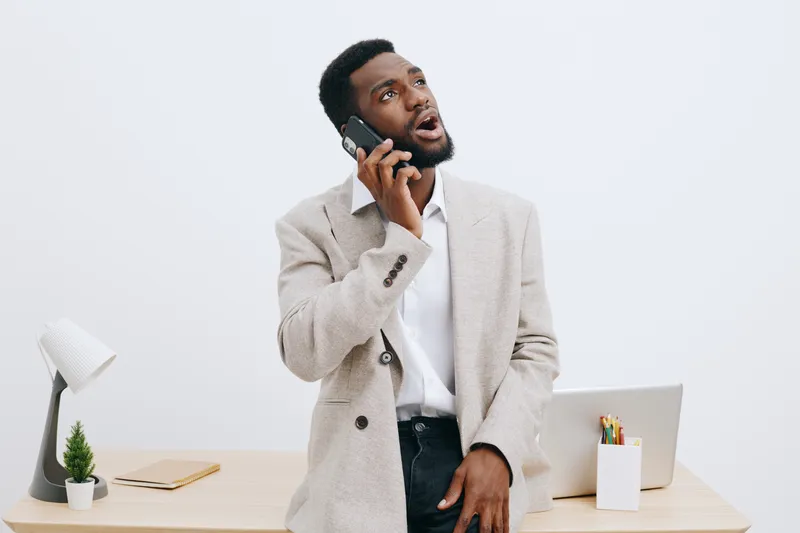 Professional young man with beard, wearing beige blazer and white shirt, talking on mobile phone, standing at modern office desk with laptop, plant, and stationery, isolated on plain light gray backgr