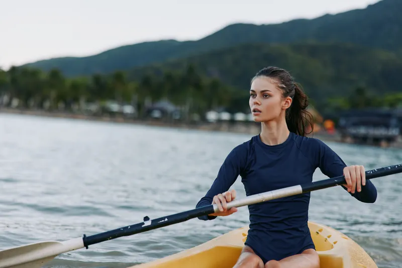 A focused young woman rows a kayak on calm water, gripping a paddle mid stroke, showcasing athletic form, concentration, and balance against a tranquil lakeside backdrop.