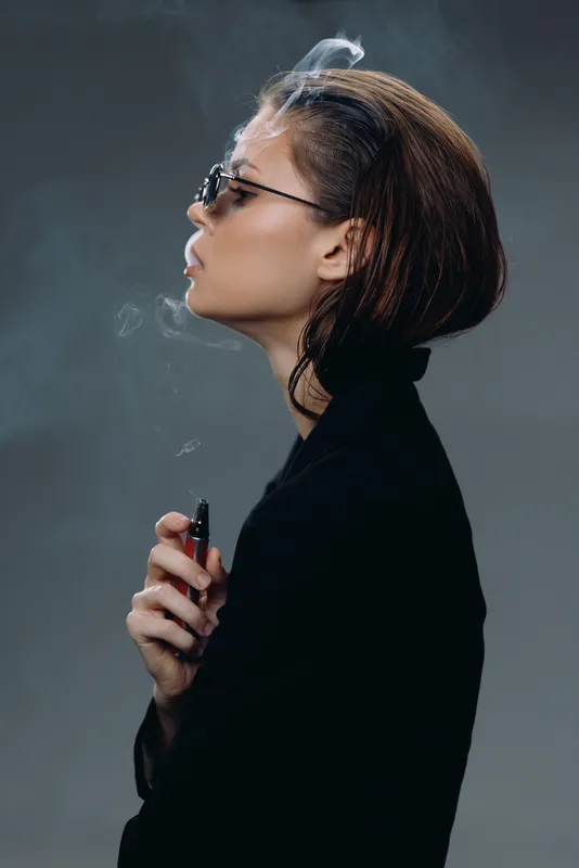 Profile of a stylish woman smoking with sunglasses and dark clothing against a gray background.