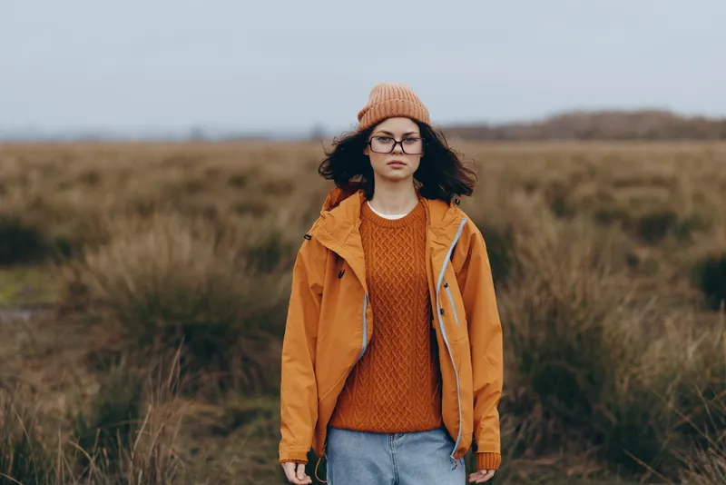 Woman lifestyle outdoor portrait in autumn field, standing woman wearing orange sweater, brown jacket, knitted hat and glasses, soft natural light, rural landscape background