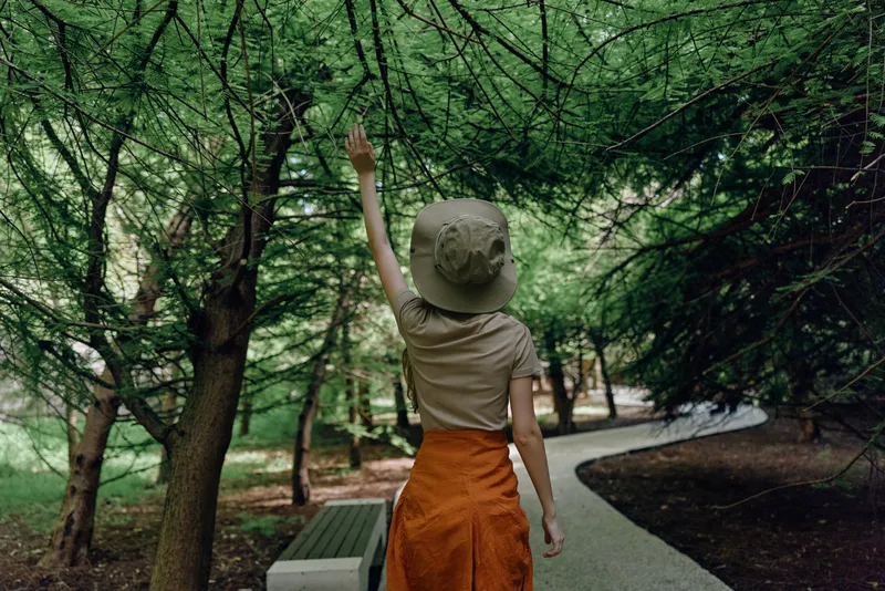 Woman in forest wearing a hat and orange skirt reaching up to tree branches on a winding path by a bench, exploring nature during a quiet summer walk in green woodland alone.