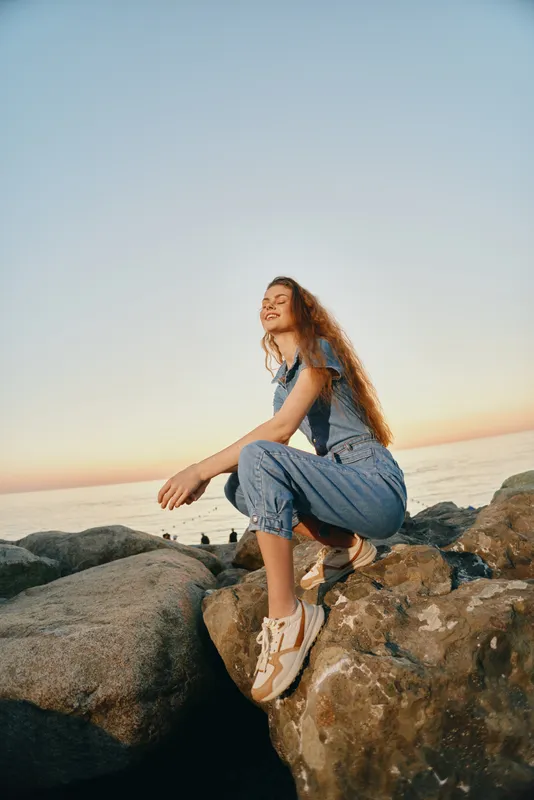 smiling woman in casual denim outfit sitting on rocks near sea during golden hour, enjoying warm sunset and fresh air with relaxed expression