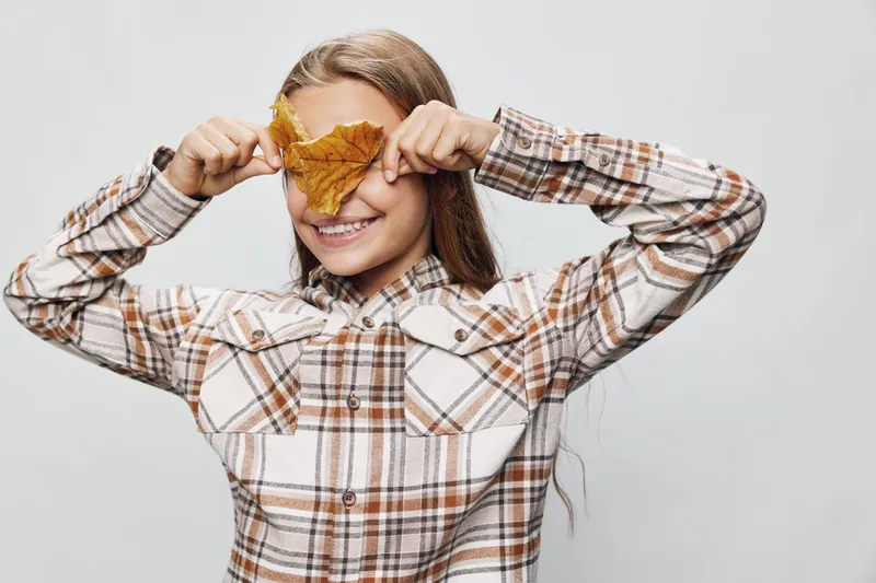 Girl smiling and covering eyes with yellow autumn leaf wearing plaid shirt standing against light background. Child having fun and playful mood with casual outfit and long hair.