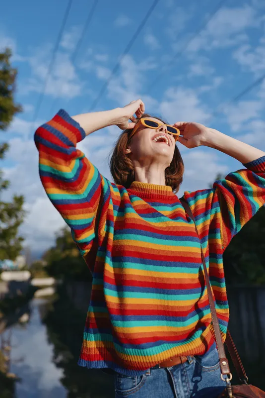 Woman in her late twenties wearing a colorful striped sweater and round sunglasses, smiling and looking up under a bright blue sky by a canal, casual outfit travel lifestyle, joyful mood.