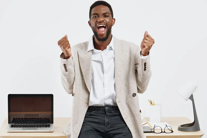 Confident young Black man in stylish beige blazer celebrating success in office, raising fists in triumph, smiling broadly, surrounded by work desk items, modern workspace, joyful mood, business achie