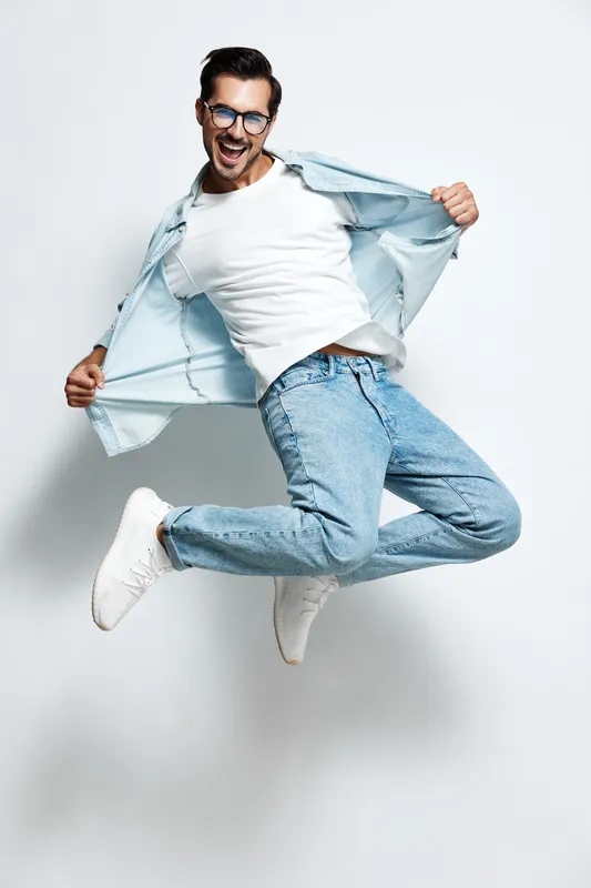 Full body joyful young man wearing casual outfit jumps in the air with a playful expression, showcasing a carefree attitude against a soft gray background. Lifestyle concept.
