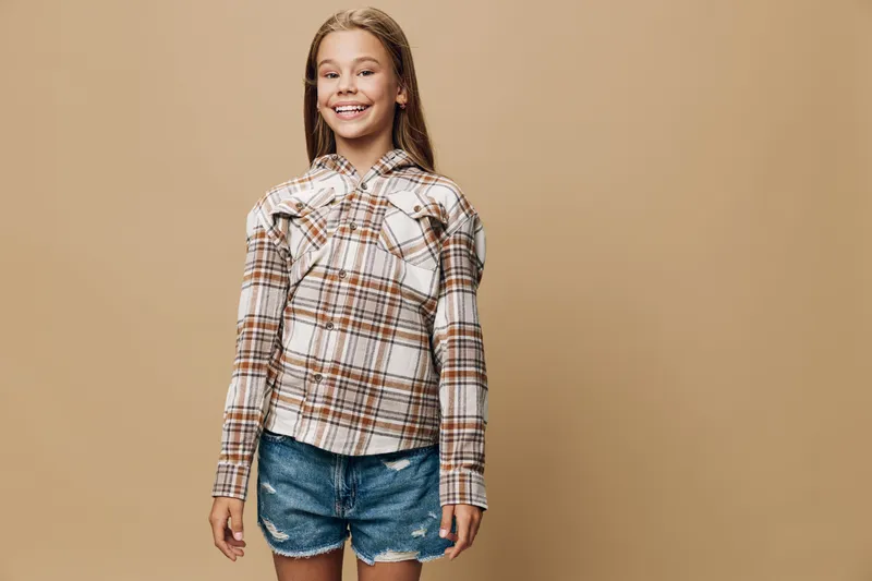 Smiling girl with long hair wearing plaid shirt and denim shorts standing against beige background. Happy child posing in casual outfit for portrait photography in studio setting.