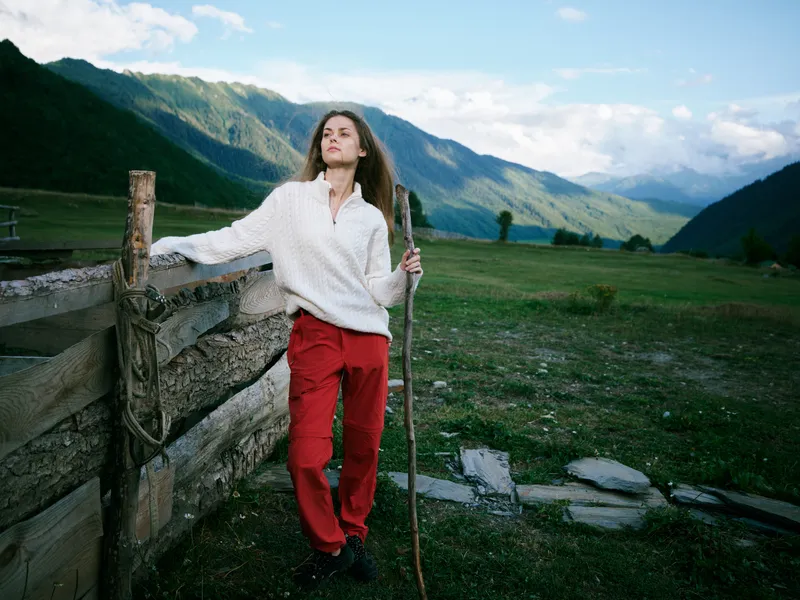 Woman hiking in a white sweater and red pants leans on a wooden fence in an expansive countryside with distant mountains, blue sky, and open landscape, embodying outdoor adventure and serenity.