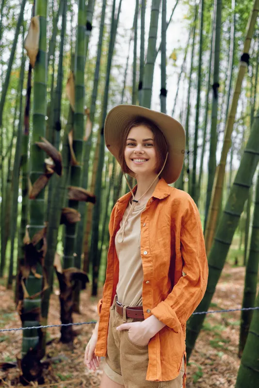 Woman smile bamboo hat orange forest portrait of a young traveler in casual clothing, straw hat and orange shirt among tall bamboo stalks, nature walk and relaxed outdoor lifestyle.