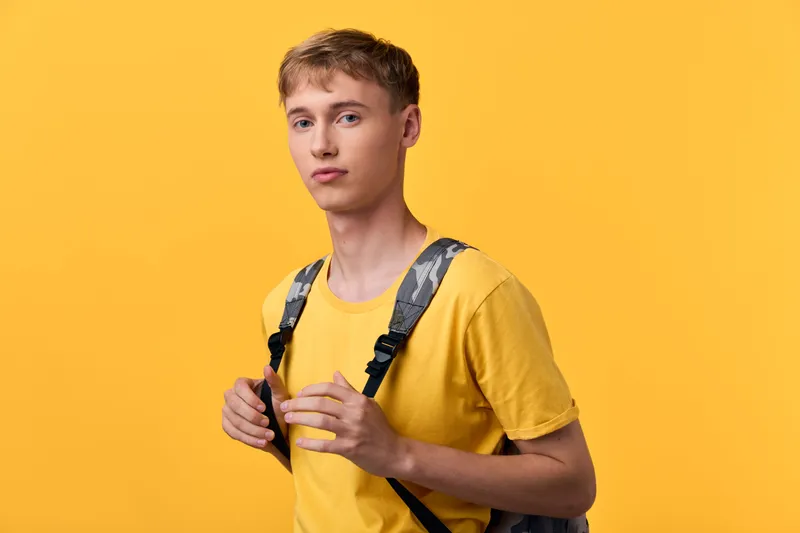 Youth wearing a yellow t shirt with a backpack, standing in studio, facing camera with a calm expression, showcasing casual fashion and everyday style for education and lifestyle content