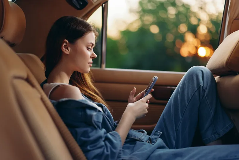 Young woman in casual denim outfit relaxing in a car, using her smartphone during sunset, evoking a sense of tranquility and modern lifestyle.