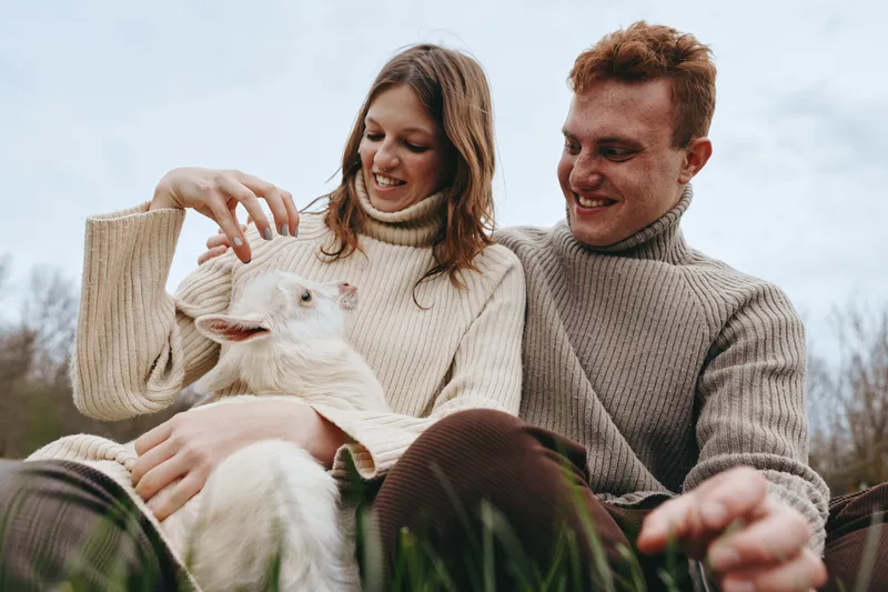 A young woman and man wearing knitted sweaters sit outdoors holding a white baby goat while smiling and enjoying a relaxed lifestyle moment in nature.